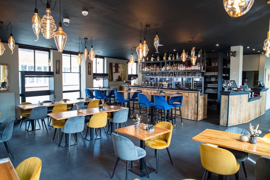 De brasserie met blauwe en okergele stoelen en de toog achteraan